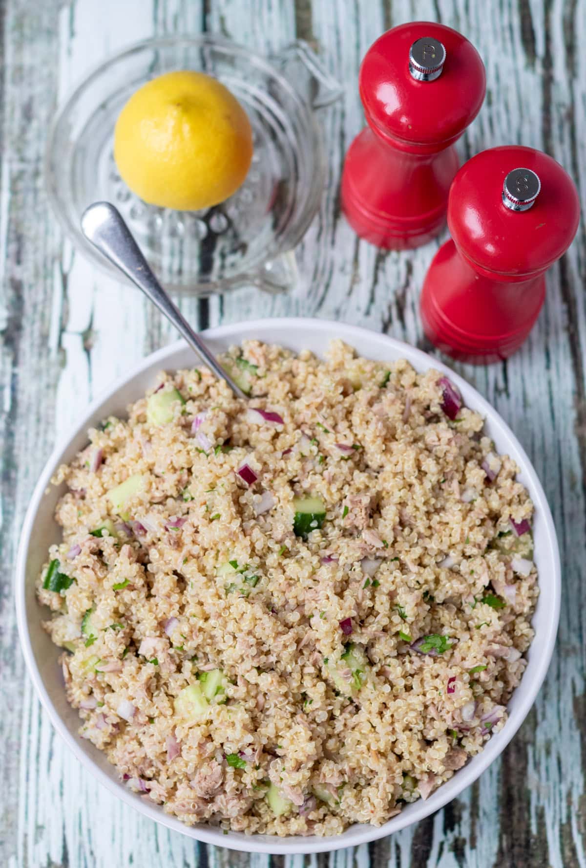 Overhead view of a serving dish of quick tuna quinoa salad with a serving spoon in. Above half a lemon in a lemon juicer and salt and pepper cellars.