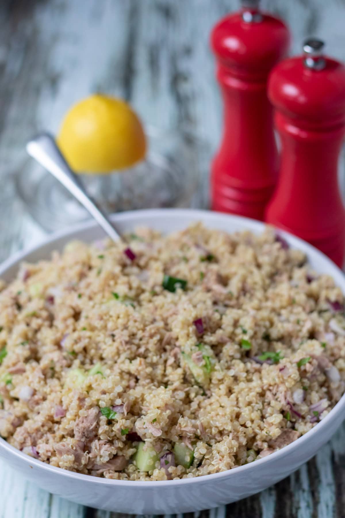 Front view of a bowl of tuna quinoa salad with a spoon in. Half a lemon in a glass lemon juicer in the background alongside salt and pepper cellars.