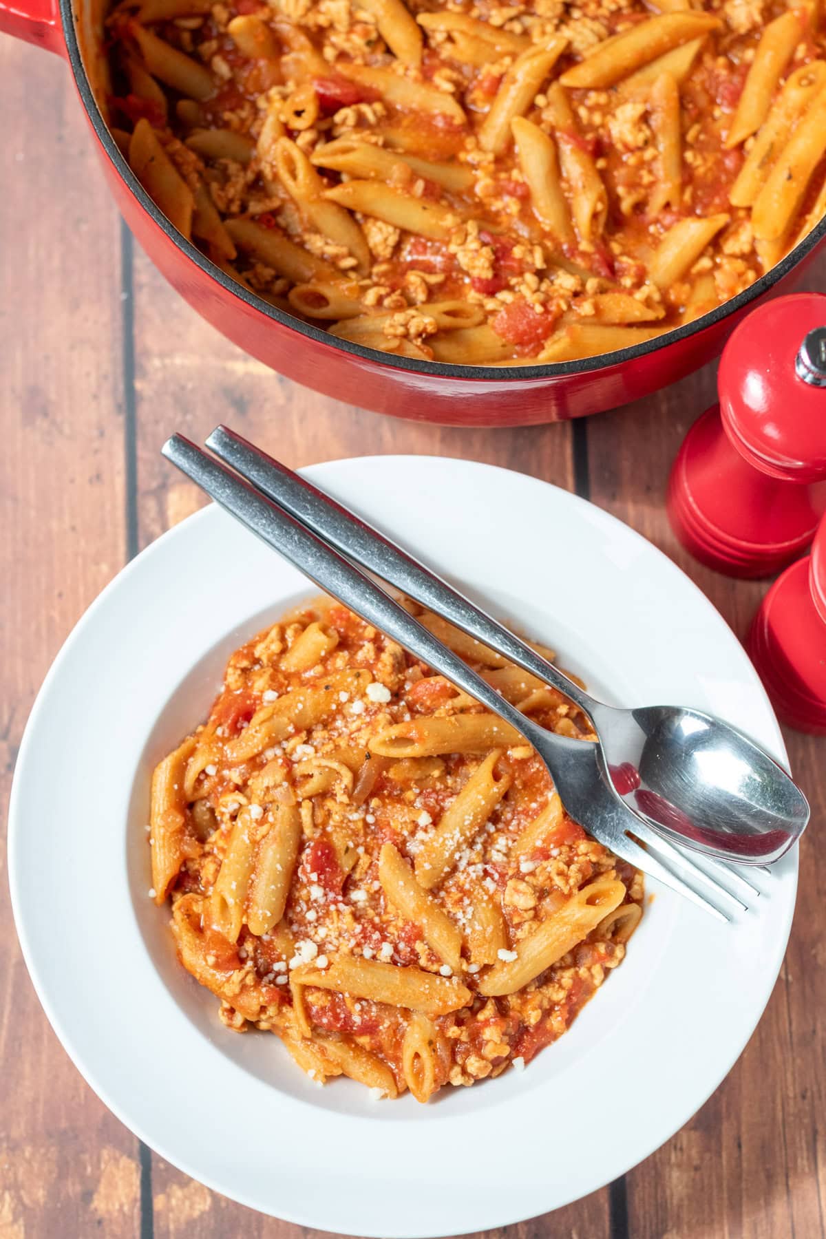 Overhead view of a bowl of turkey mince pasta with a spoon and fork on the side. Above is the rest of the recipe in a large casserole pot and salt and pepper cellars.
