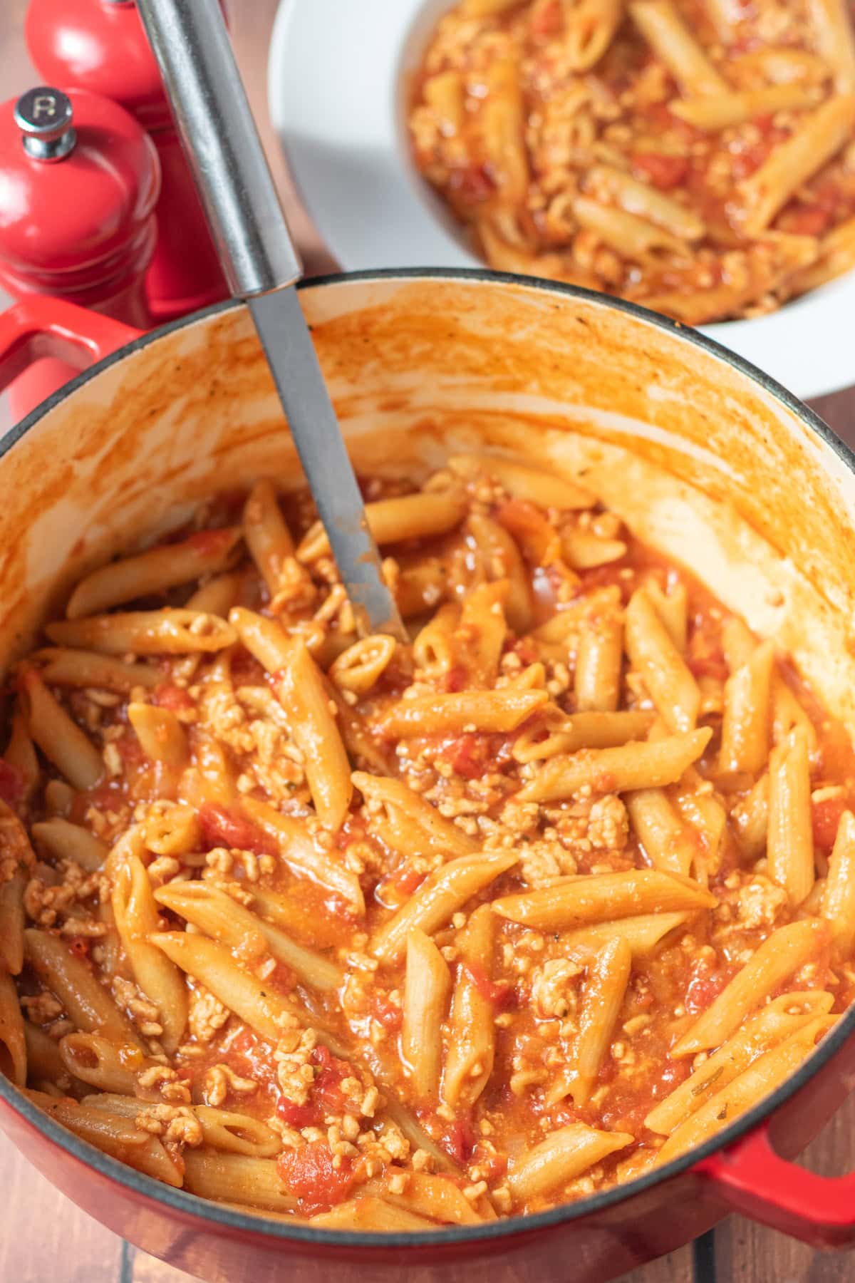 One pot turkey mince pasta in a large casserole pot with a serving spoon in. Behind is a portion served in a pasta bowl with salt and pepper cellars alongside.