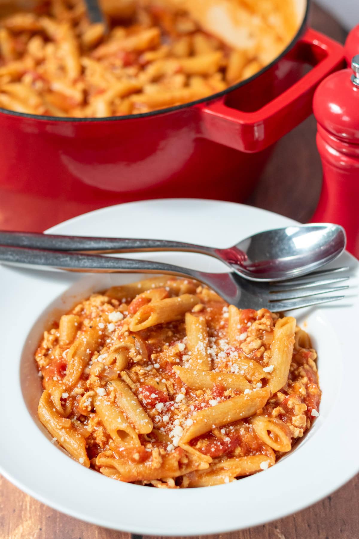A serving of turkey mince pasta in a pasta bowl with a fork and spoon on the rim. Rest of the recipe in casserole pot above.
