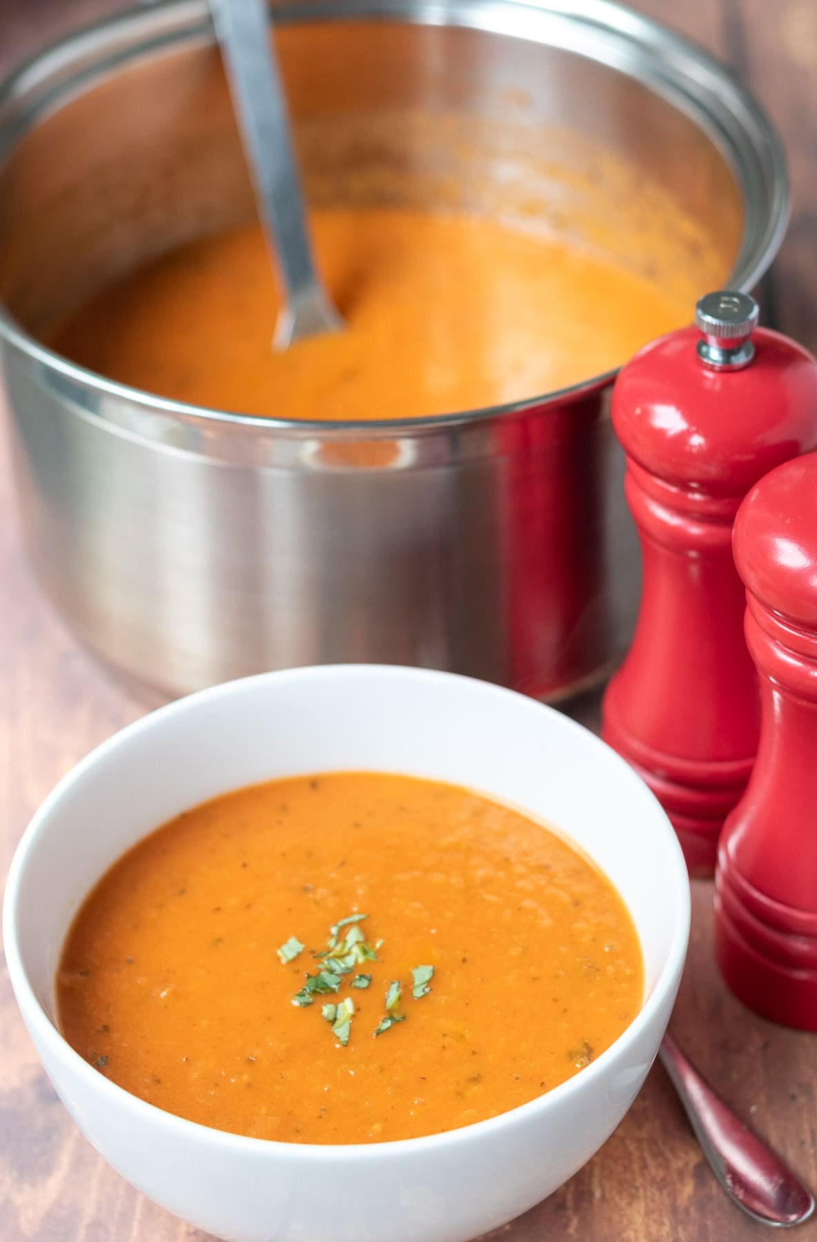 Close up view of a bowl of roasted tomato and basil soup. To the right salt and pepper cellars and behind a large saucepan with the remainder soup in and a ladle.