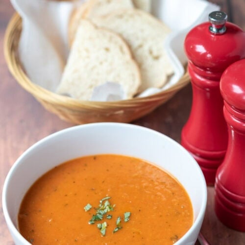 A bowl of simple roasted tomato basil soup garnished with freshly chopped basil. To the right salt and pepper cellars at the back a basked of sliced bread.