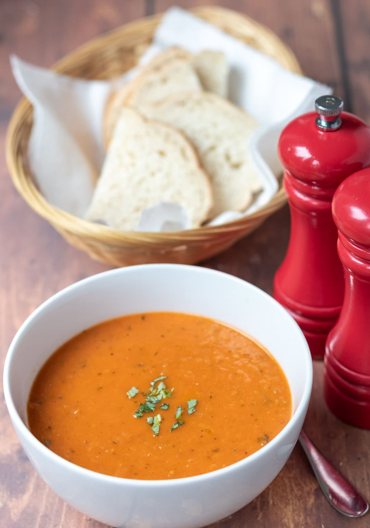 A bowl of simple roasted tomato basil soup garnished with freshly chopped basil. To the right salt and pepper cellars at the back a basked of sliced bread.