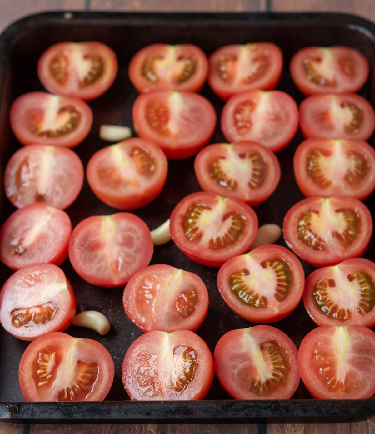 Tomatoes cut side up on a large baking tray with 4 garlic cloves sprinkled amongst the tomatoes.