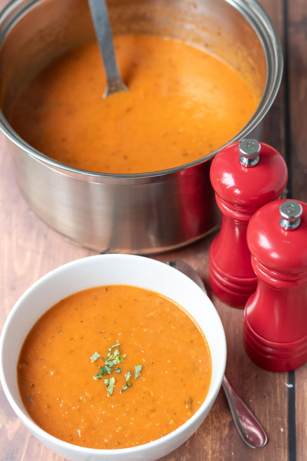 Bowl of simple roasted tomato basil soup garnished with chopped basil at the front. Salt and pepper cellers to the right and the remaining soup in the large saucepan at the back with a soup ladle in.