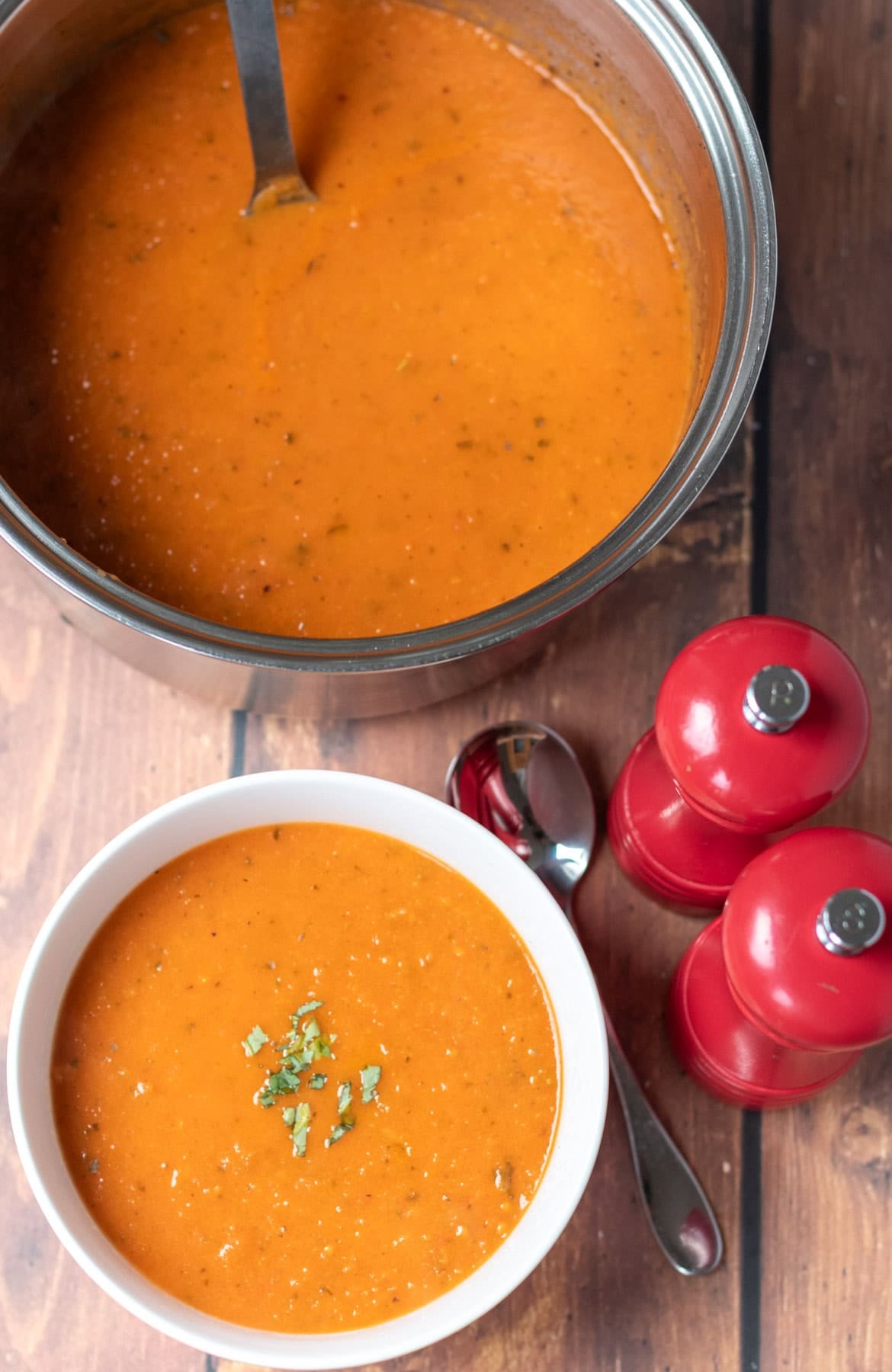 Overhead view at the bottom a bowl of roasted tomato basil soup garnished with chopped basil, and a soup spoon and salt and pepper cellars to the right. Above the rest of the soup in a large saucepan with a soup ladle in.
